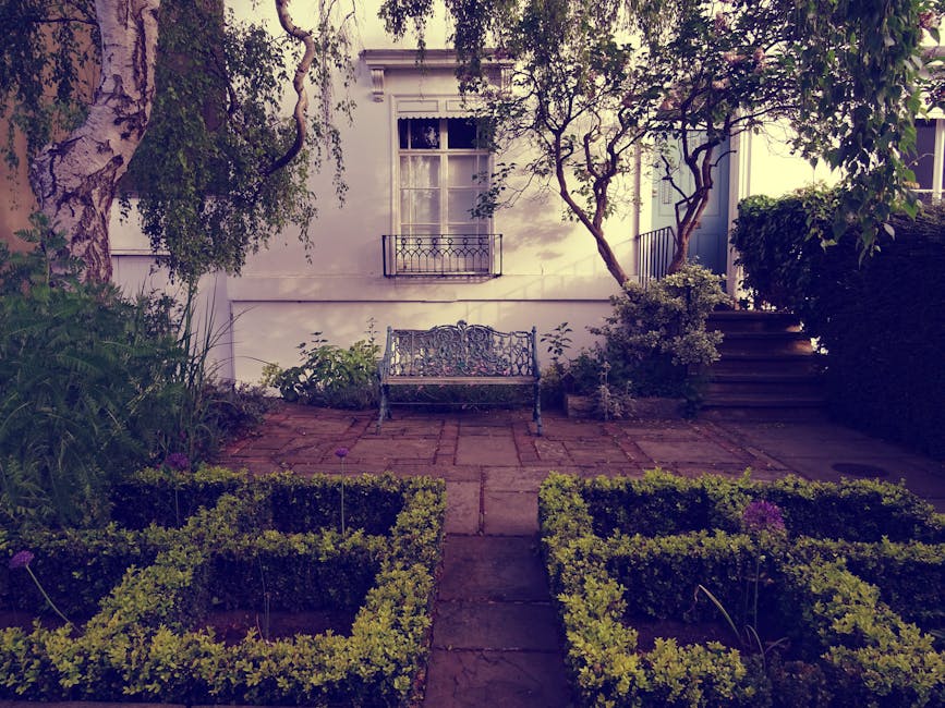 A neatly maintained courtyard garden featuring a lush green lawn bordered by low hedges and flower beds with fresh, trimmed foliage. The garden has a paved pathway with a natural stone surface winding